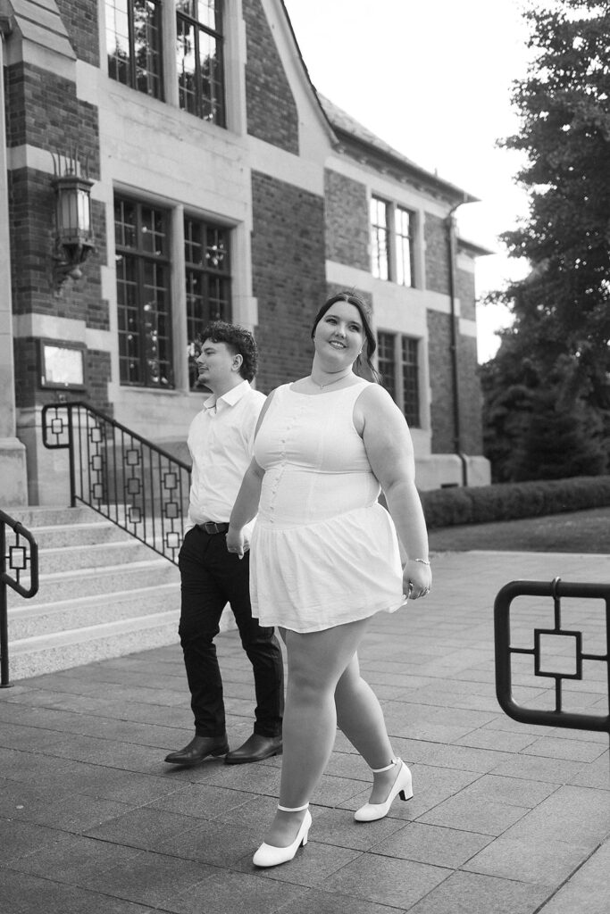 A woman in a white dress and heels walks in front of a man in a white shirt and dark pants outside a brick building with large windows, both appearing happy and relaxed.