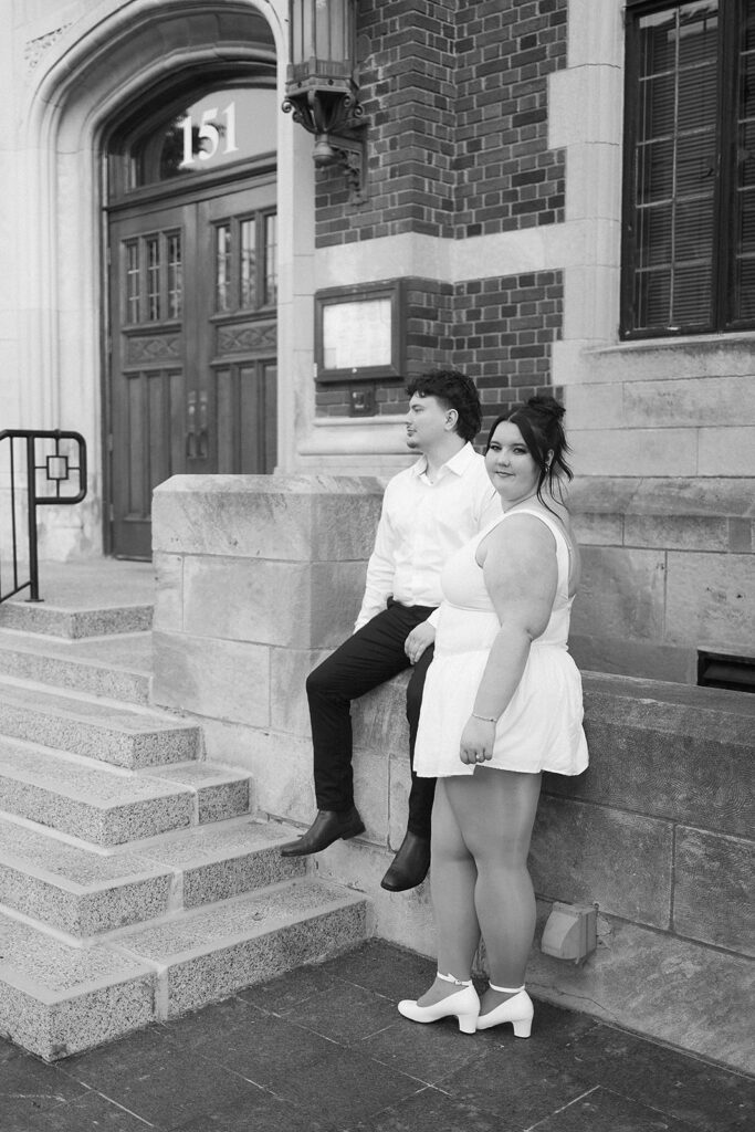 A couple poses outside a brick building with the address 151. The woman in front wears a white dress and heels, while the man behind her sits on a ledge in a white shirt and dark pants. Both look in different directions.