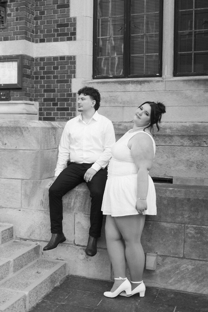 A man in a white shirt and dark pants sits on a stone ledge, while a woman in a white dress and ankle boots stands beside him, smiling. They are outside a brick building with large windows.