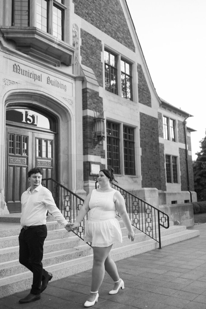 A couple holding hands walks down the steps of a brick Municipal Building. The man looks back at the woman, who is wearing a white dress and heels. Both appear happy and engaged with each other.