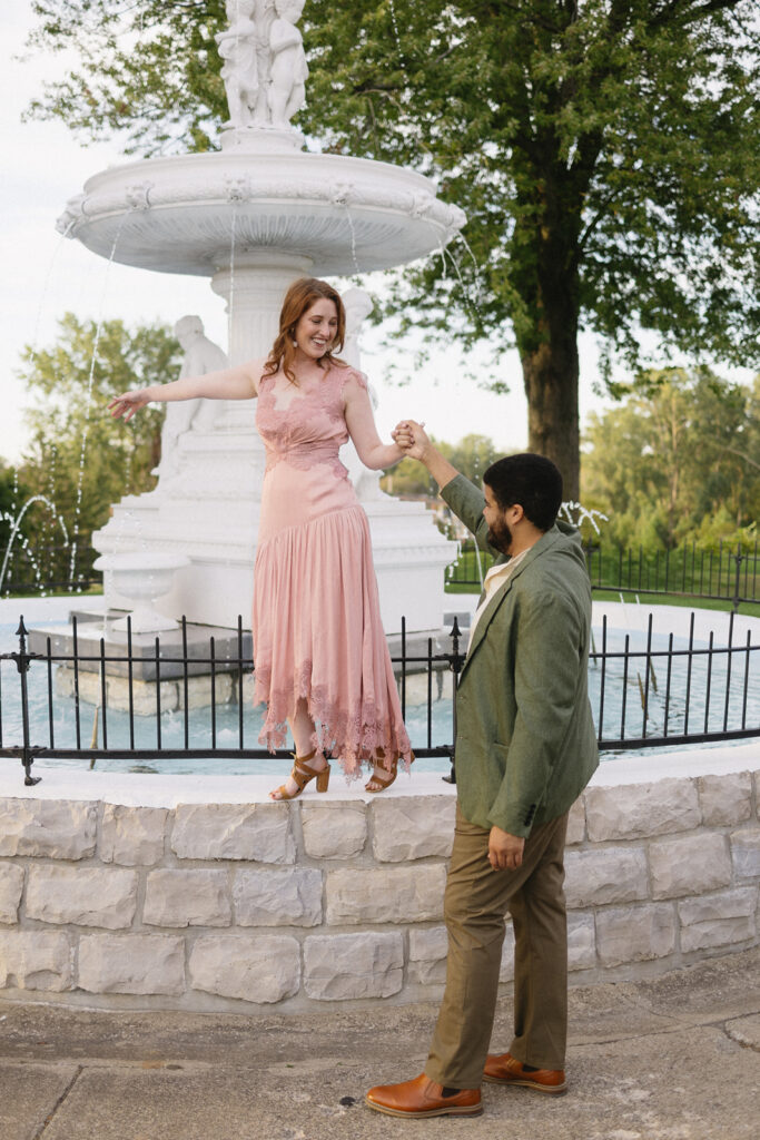 A woman in a pink dress balances on the edge of a stone fountain, holding hands with a man in a green jacket who stands beside her, both smiling. Water sprays from the white fountain in the background.