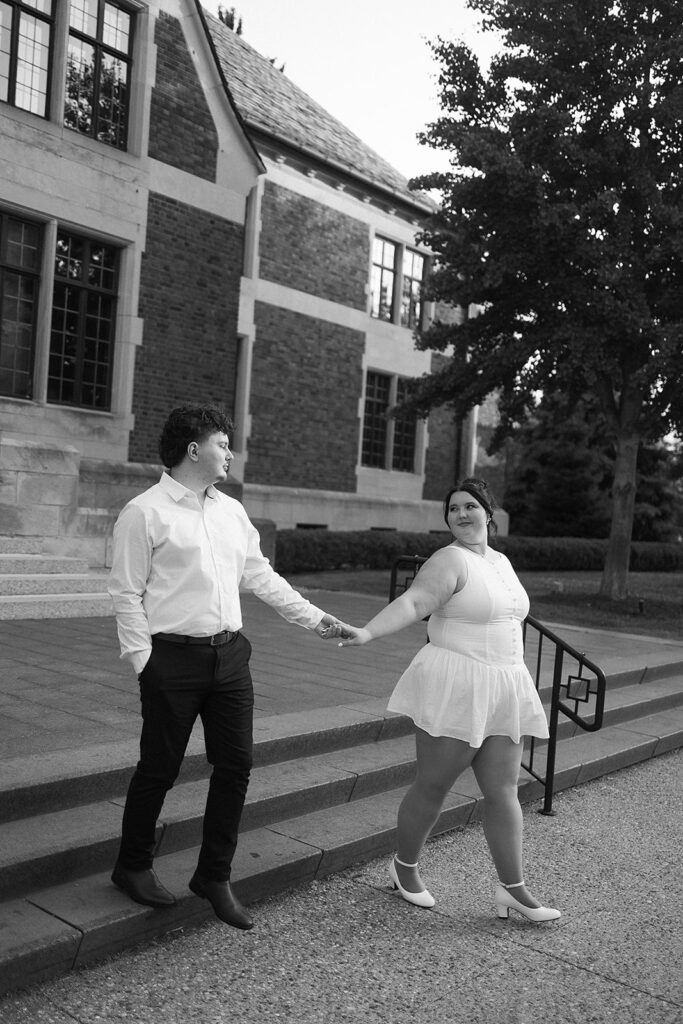 A couple holds hands while walking outside a brick building. The woman, in a short white dress and boots, looks back at the man, who wears a white shirt and dark pants. They share a moment on a paved walkway.