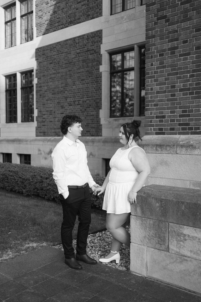 A couple stands outside a brick building, holding hands and looking at each other. The man wears a white shirt and dark pants, while the woman wears a white sleeveless dress. The scene is in black and white.