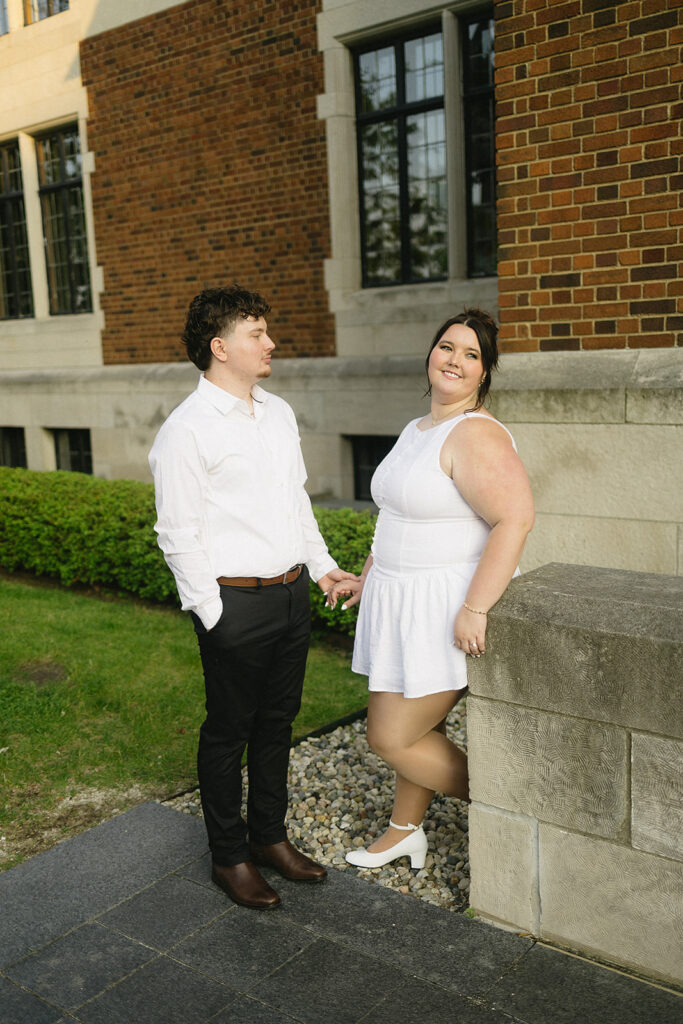 A couple stands outside a brick building. The man wears a white shirt and black pants; the woman wears a white dress and heels, leaning against a stone ledge. They are holding hands and looking at each other, smiling.