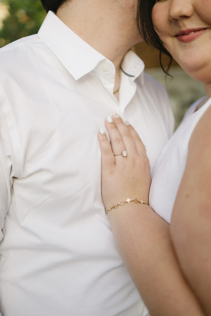 A close-up of a couple embracing, focusing on the woman’s hand with a ring resting on the mans chest. Both wear white outfits; her nails are painted white with black dots, and she wears a gold bracelet. Their faces are partially visible.