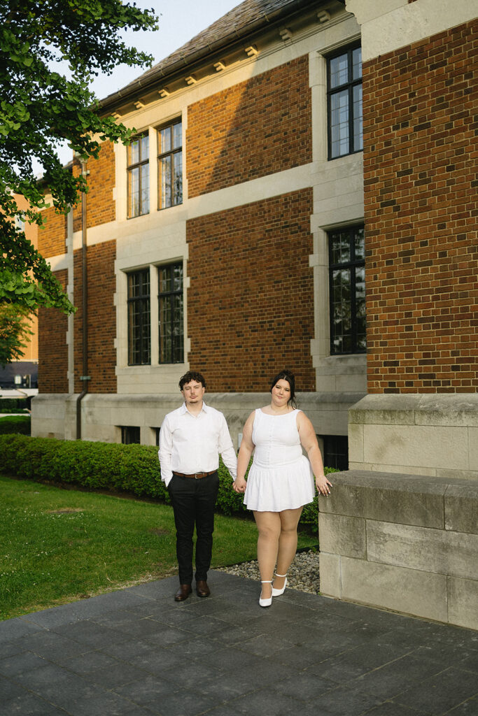 A couple stands holding hands outside a large brick building with tall windows. Both wear white outfits; the man wears black pants, while the woman wears a short dress and flats. Green grass and a hedge line the walkway beside them.