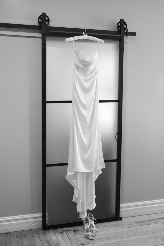 A white, sleeveless wedding dress hangs on a sliding frosted glass door. A pair of strappy high-heeled sandals is placed below the dress on a wooden floor. The image is in black and white.