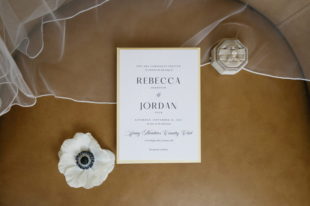 Wedding invitation on a tan surface with a white flower, a sheer white veil, and a light-colored ring box with rings inside. The invitation is addressed to Rebecca and Jordan for a September 26, 2025 wedding.