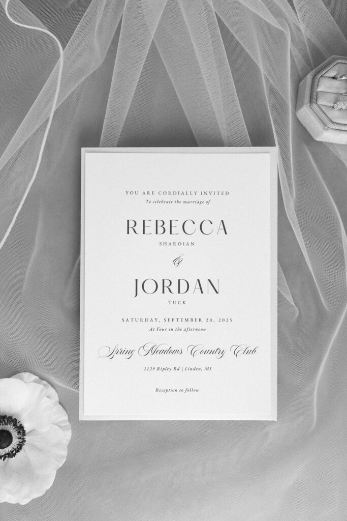 A wedding invitation on white paper atop sheer fabric, featuring black elegant text for Rebecca Sharolan and Jordan Tuck’s ceremony on September 20, 2025, at Spring Meadows Country Club. A white flower and ring box are nearby.