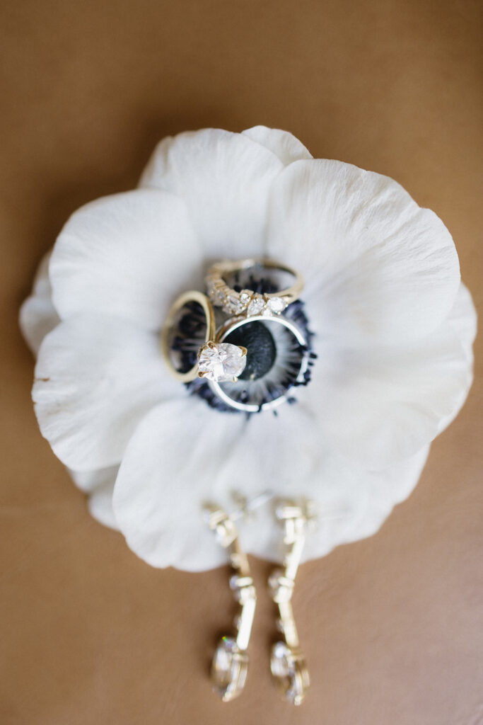 Gold rings with diamonds rest on the center of a white flower, with blurred gold earrings placed below, all set against a light brown background.