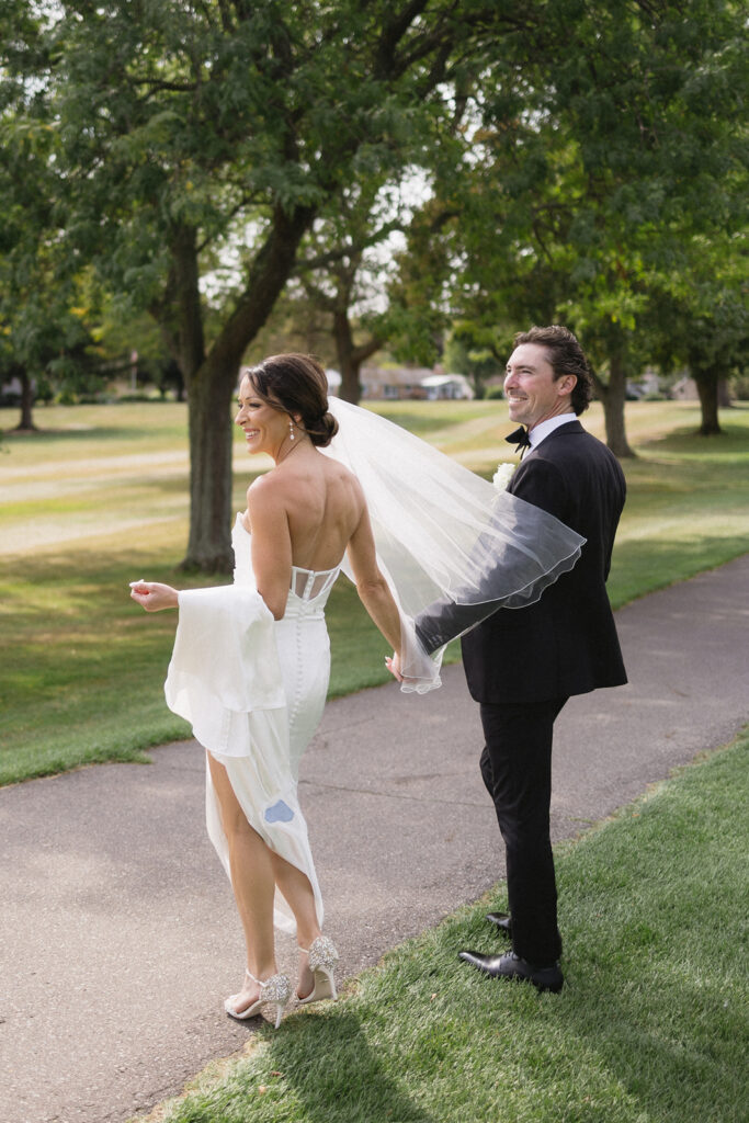 A bride in a white dress and veil holds hands with a groom in a black suit as they walk together on a tree-lined path in a park, both smiling and looking back over their shoulders.