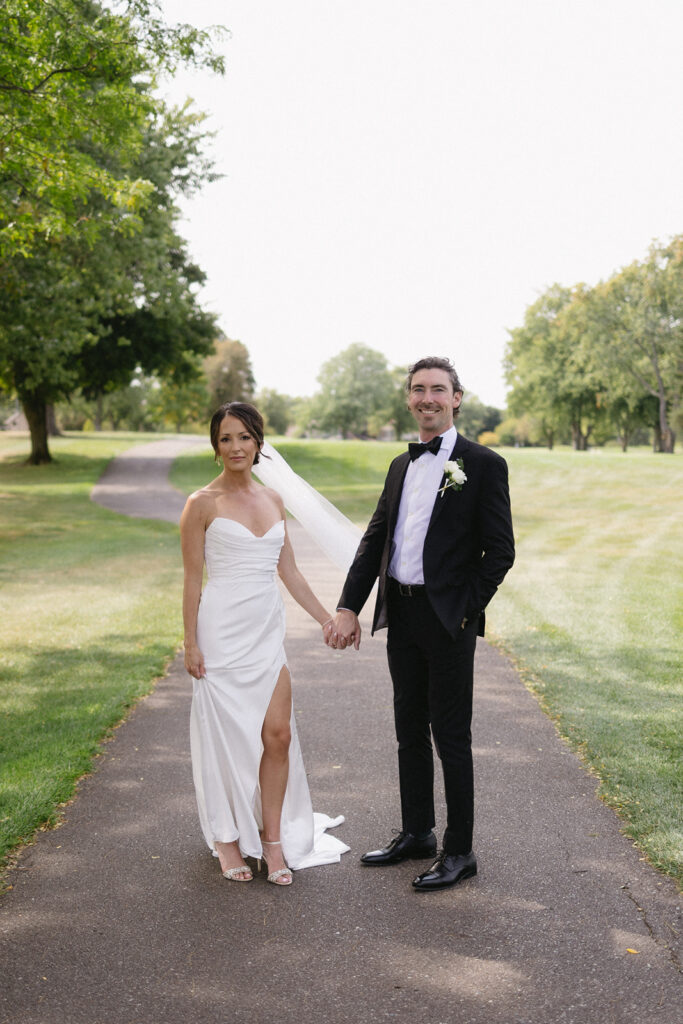 A bride in a white dress and a groom in a black suit stand holding hands on a paved path in a grassy park, surrounded by trees, both facing the camera and smiling softly.