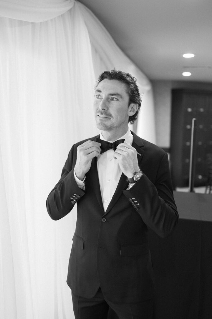 A man in a dark suit and bow tie adjusts his collar while standing indoors near white draped curtains. The setting appears formal, and the image is in black and white.