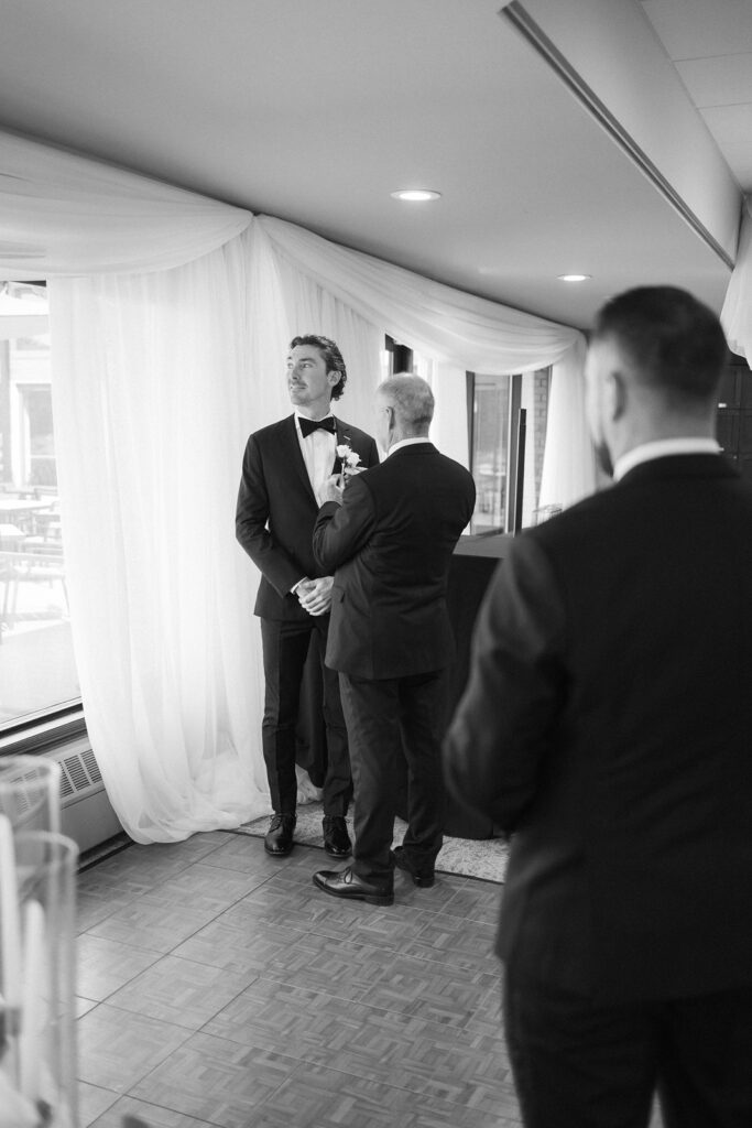 A groom in a tuxedo stands near a window with draped white curtains, looking upward and smiling as an older man adjusts his boutonnière. Another man in a suit stands in the foreground facing them.