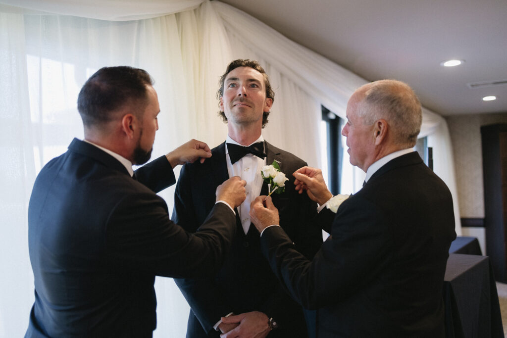 Three men in suits adjust a grooms boutonniere and bow tie in a softly lit room with white drapes. The groom stands in the center, looking slightly upward with a calm expression.