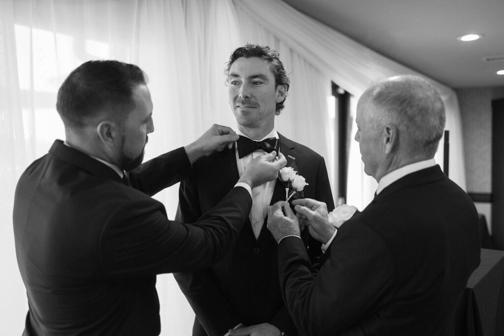 A groom stands smiling while two men adjust his bow tie and boutonnière, preparing him for a wedding. All three are dressed in suits, and the scene takes place indoors with soft natural light.